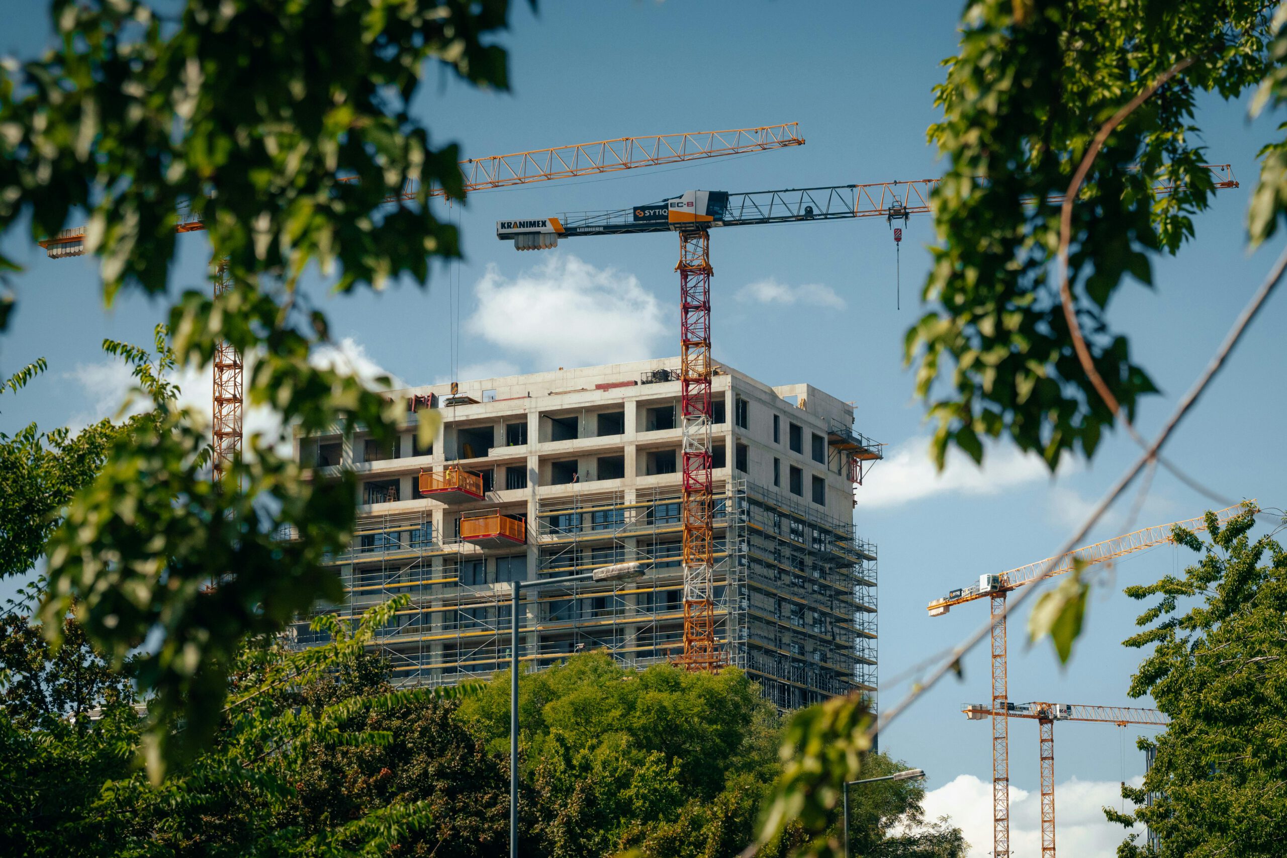 High-rise building under construction with cranes in Bratislava, seen through lush green trees.